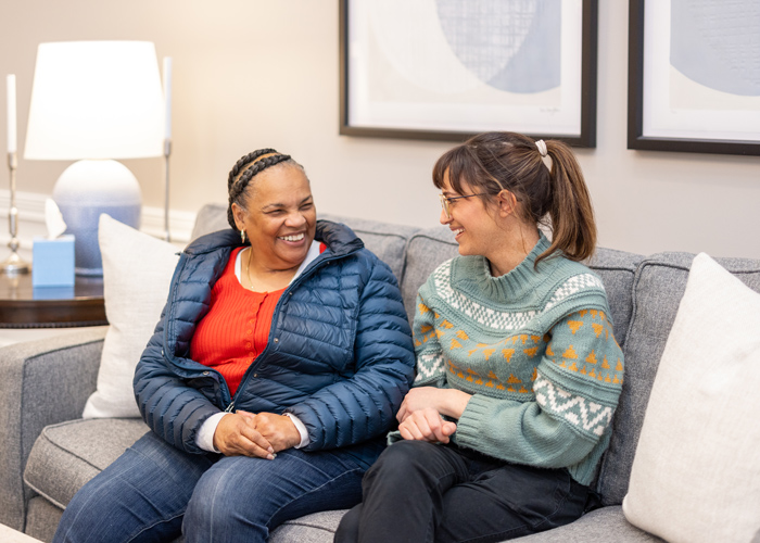 Two women, one in a blue jacket and the other in a green sweater, share a warm, joyful conversation while sitting on a comfortable gray couch in a cozy senior living community.