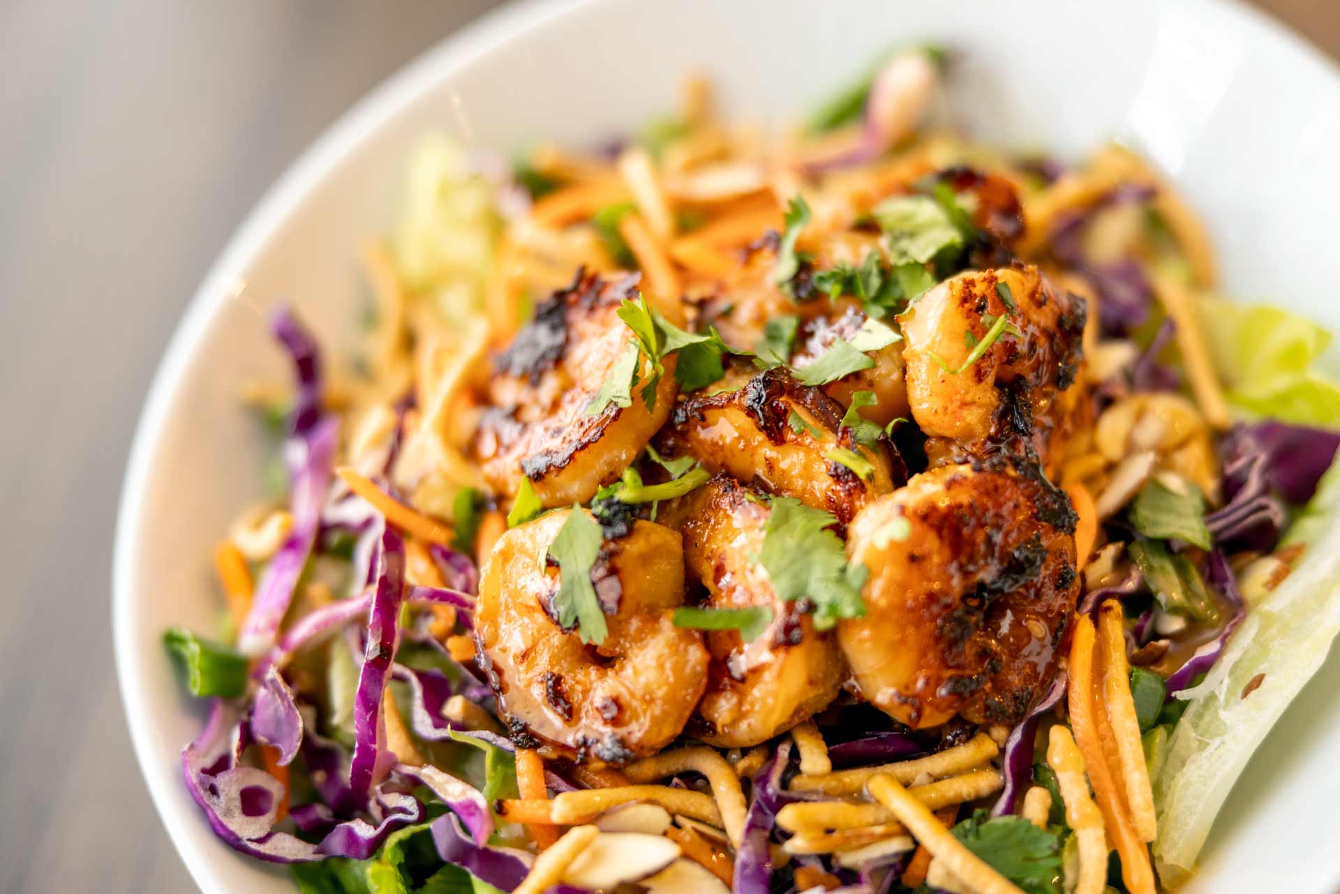 Close-up of a vibrant dish featuring grilled shrimp garnished with cilantro, served on a bed of fresh shredded vegetables including cabbage and carrots, topped with herbs.
