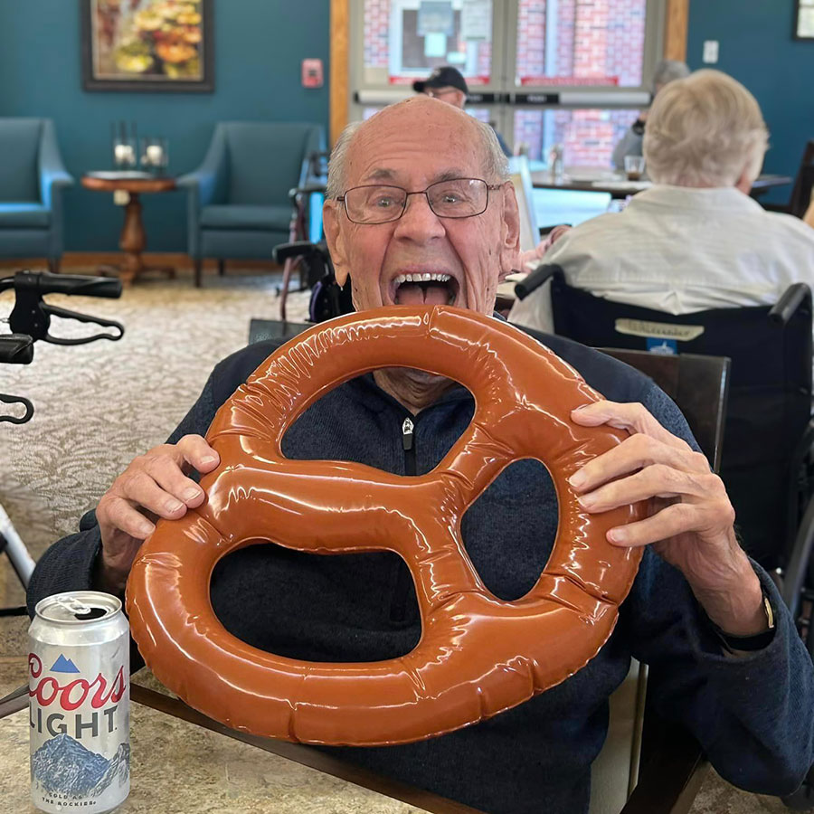 Smiling senior resident holds up a large inflatable pretzel, playfully pretending to take a bite, with a can of Coors Light beer on the table in front of him, celebrating National Pretzel Day.